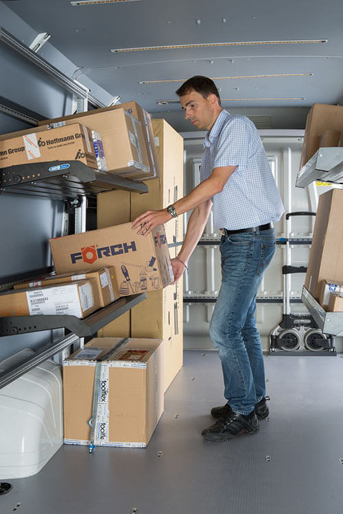 Innenraum eines Transporters. Regalsystem mit Paketen an der linken Innenwand. Person fässt ein Paket an